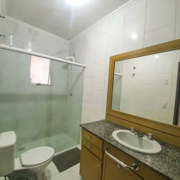 Banheiro com box de vidro, vaso sanitário, pia com bancada de granito, gabinete de madeira e espelho grande.