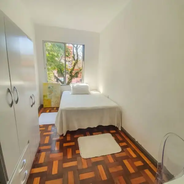 Quarto com cama de lençol branco, guarda-roupa branco, tapete pequeno e janela com árvores lá fora.