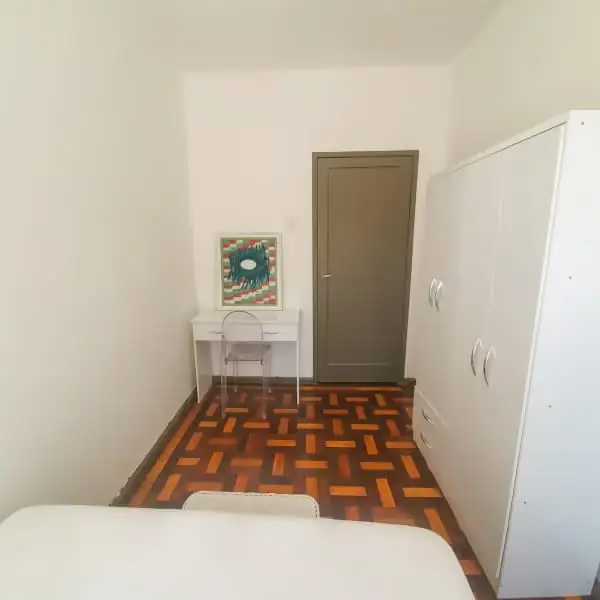 Quarto com guarda-roupa branco, mesinha branca com cadeira transparente e quadro, piso de taco e a cama em primeiro plano.