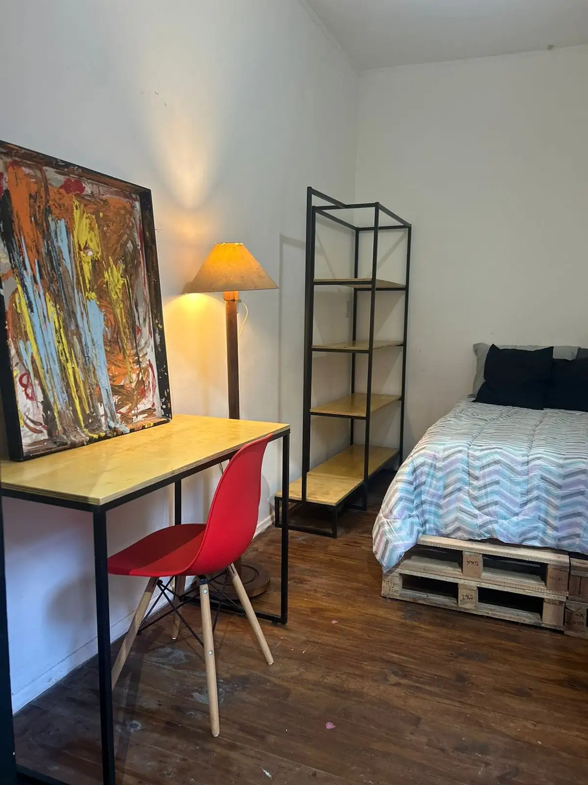 Quarto com cama sobre pallets, estante de metal e madeira, luminária de pé, mesa com cadeira vermelha e quadro colorido.