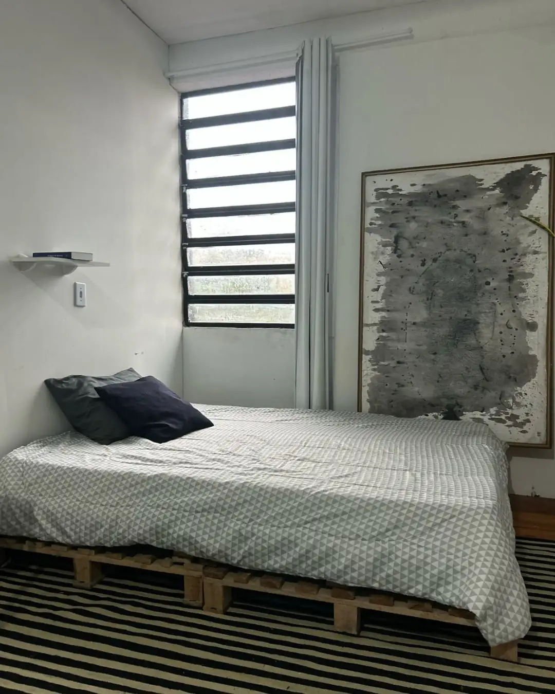 Quarto com cama sobre pallets e almofadas pretas, janela com cortina, quadro grande em tons de cinza e tapete listrado.