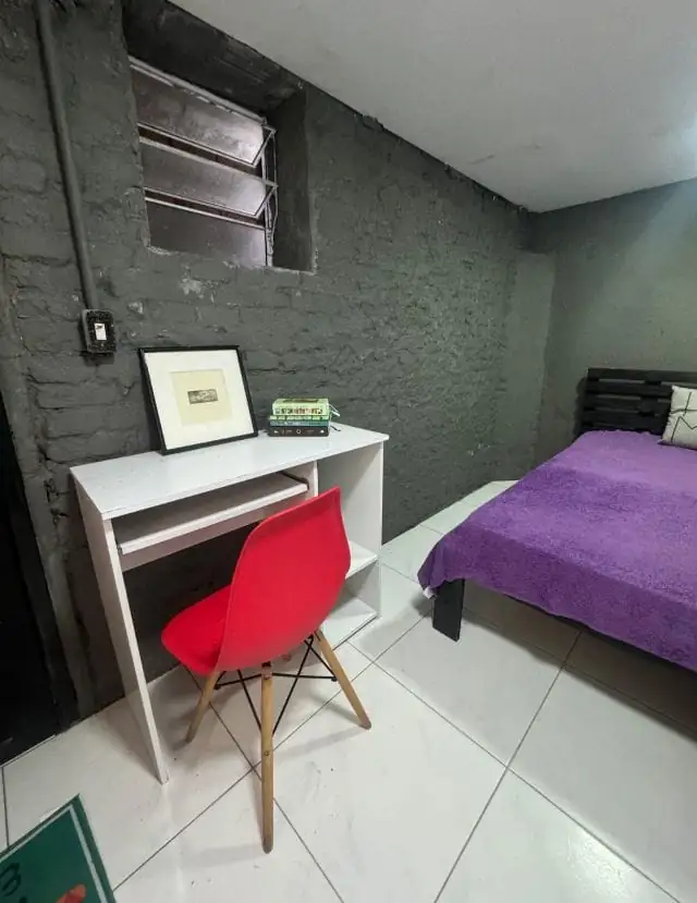 Quarto com cama de colcha roxa, escrivaninha branca com cadeira vermelha e quadro, e parede de tijolo pintada de cinza.