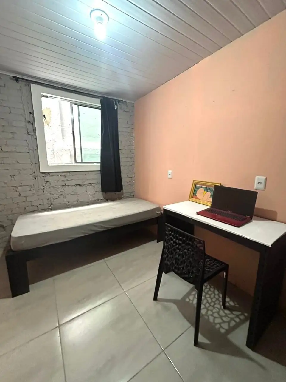 Quarto com cama de solteiro, mesa branca com cadeira preta, notebook e quadro pequeno, parede rosada e janela com cortina preta.