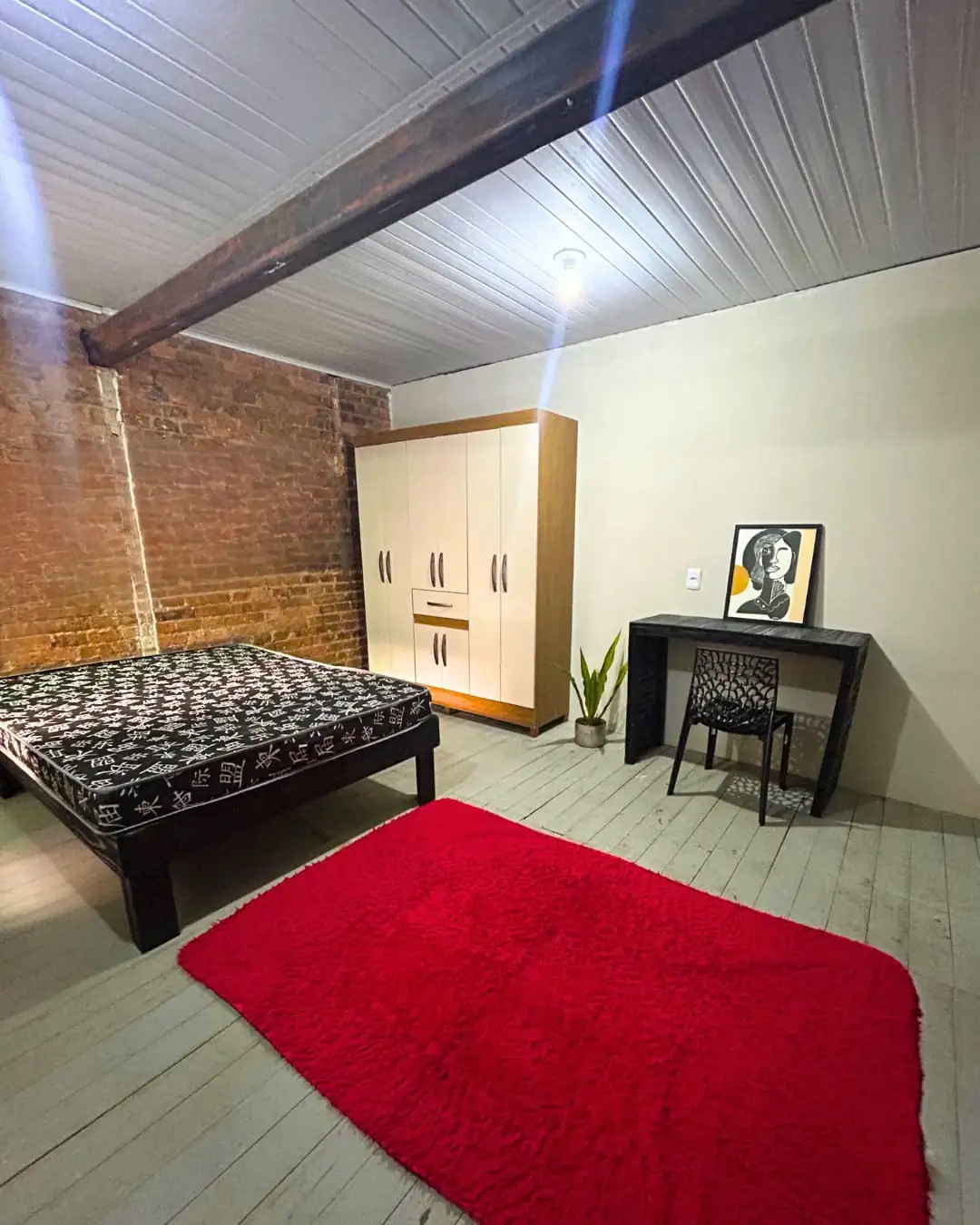 Quarto com cama de colcha estampada, tapete vermelho, guarda-roupa claro, mesa com cadeira, viga de madeira no teto e parede de tijolo à vista.