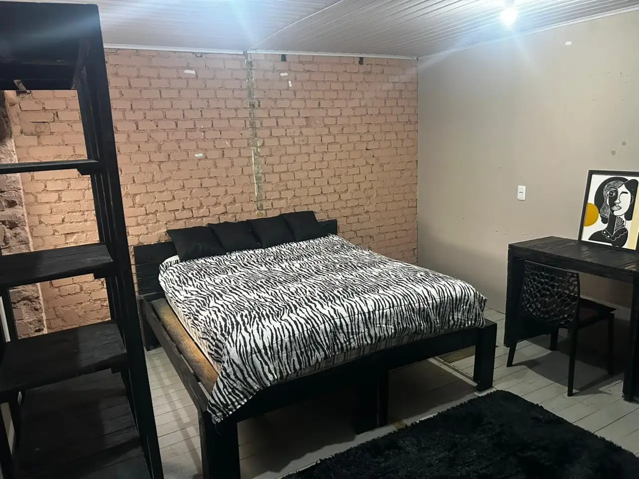 Quarto com cama de casal de colcha zebrada, parede de tijolo à vista, estante preta, mesa com cadeira e tapete preto.