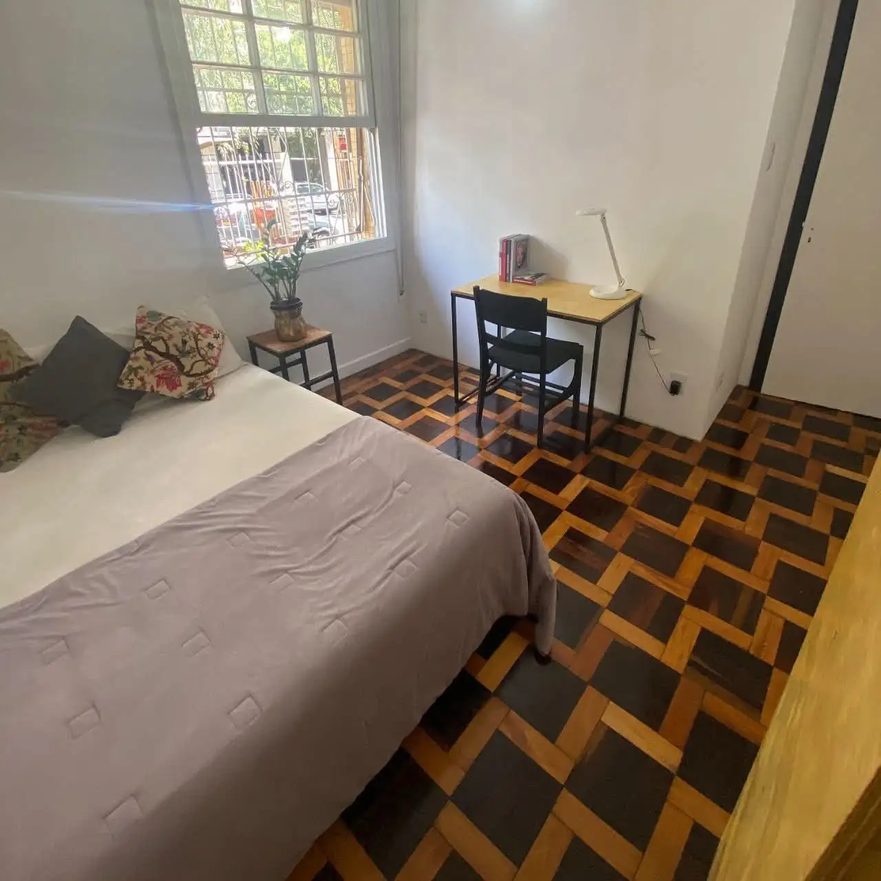 Quarto com cama e almofadas estampadas, mesa com cadeira e luminária perto da janela, banqueta com planta e piso de taco.