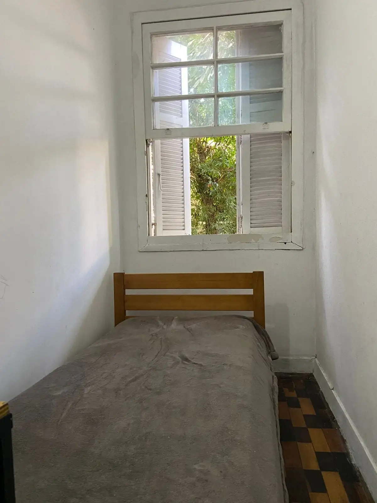 Quarto com cama de solteiro de madeira encostada na parede, sob uma janela com venezianas abertas e árvores lá fora.