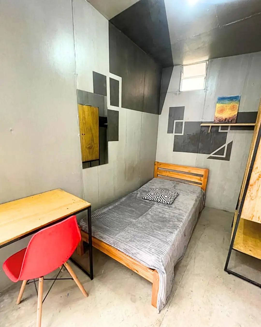 Quarto com cama de solteiro de madeira, mesa de madeira com cadeira vermelha, armário de madeira e parede com desenhos geométricos em cinza.
