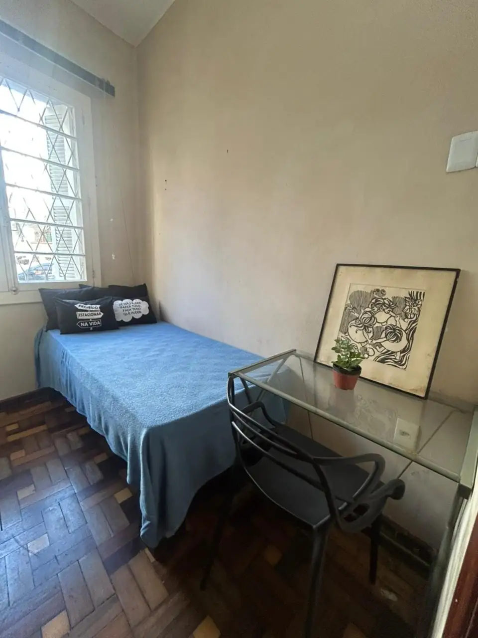 Quarto com cama de solteiro de colcha azul e almofadas, mesa de vidro com cadeira preta e planta, quadro apoiado na parede e janela com grade em losango.