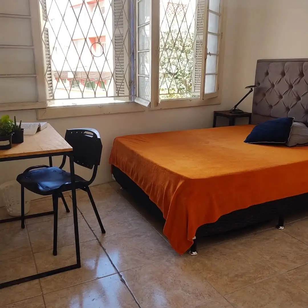 Quarto com cama de casal de colcha laranja e cabeceira estofada cinza, mesa de madeira com cadeira e janela com grade em losango.