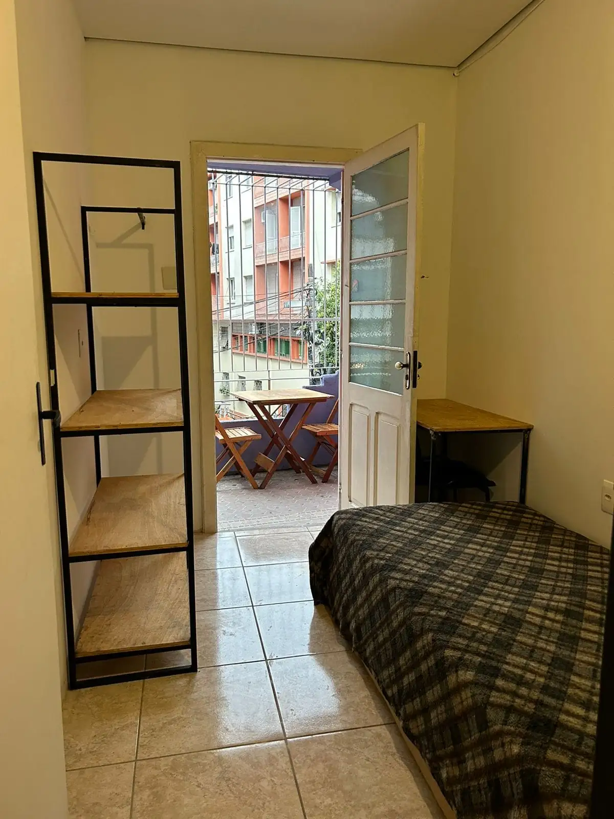 Quarto com cama de colcha xadrez, estante de metal e madeira, mesa pequena e porta de vidro aberta para uma área externa com mesa e cadeiras.