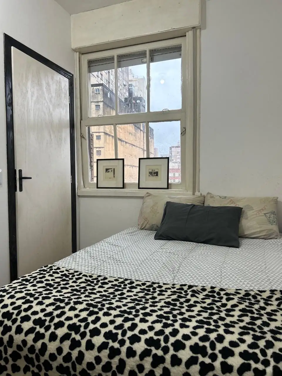 Quarto com cama de colcha estampada em preto e branco, almofadas, dois quadros pequenos no parapeito e janela com prédios lá fora.