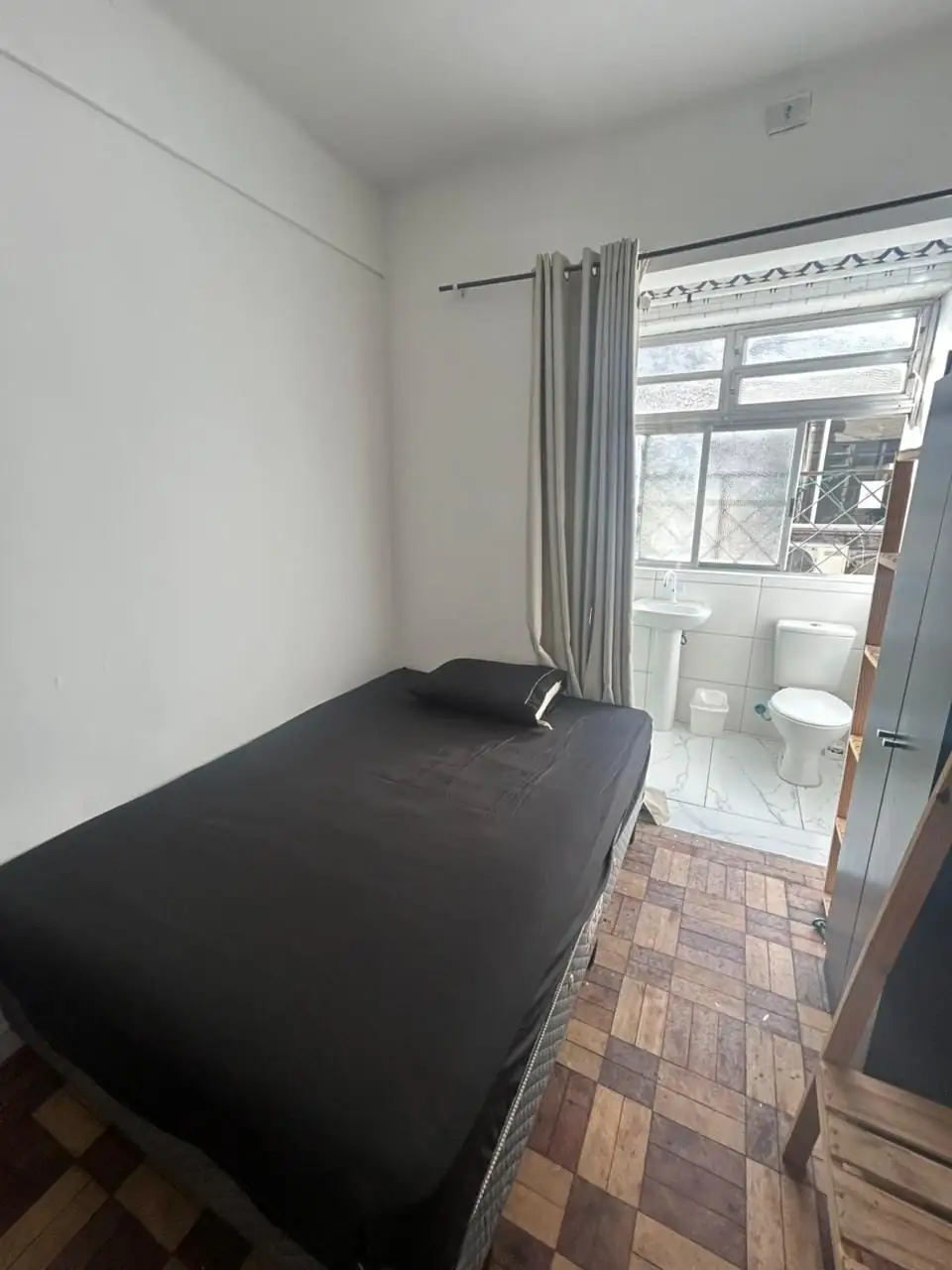 Suíte com cama de lençol preto, cortina cinza e, ao fundo, o banheiro com pia e vaso sanitário.