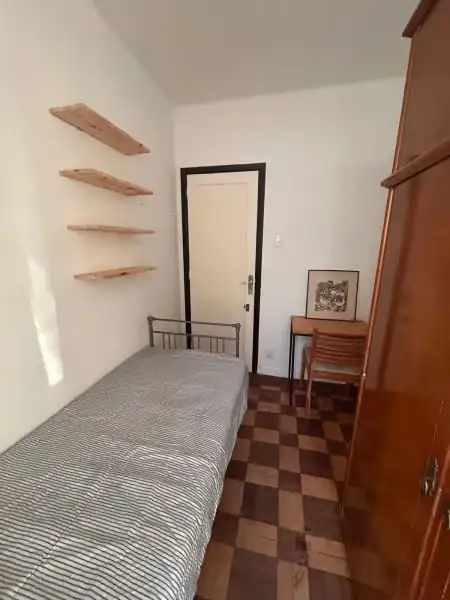 Suíte com cama de solteiro de metal, prateleiras de madeira na parede, cadeira, quadro pequeno e armário de madeira.