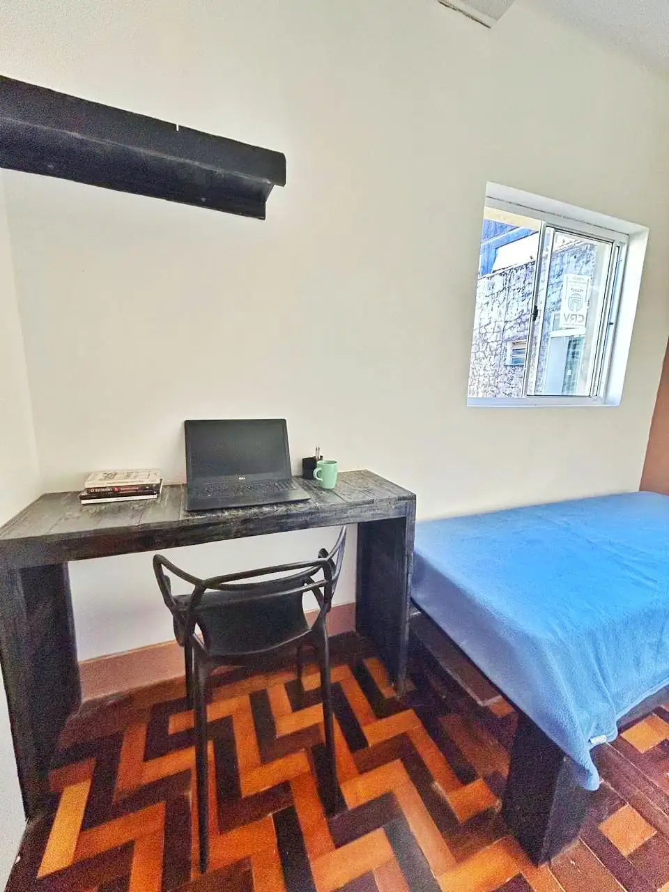 Quarto com cama de solteiro de colcha azul, mesa preta com cadeira, notebook e livros, janela e piso de taco em zigue-zague.