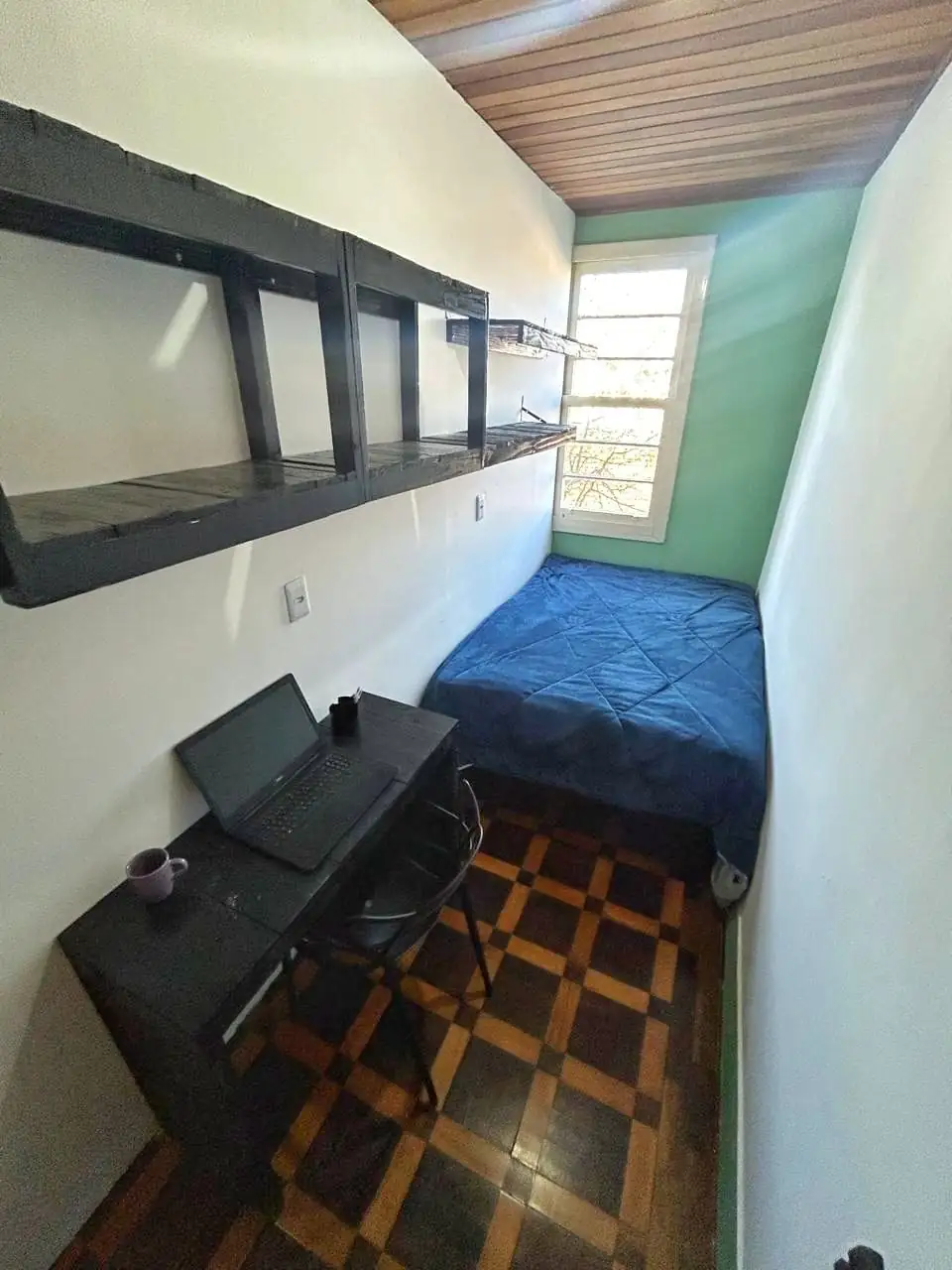 Quarto com cama de solteiro de colcha azul, mesa preta com notebook e cadeira, prateleiras pretas na parede, parede verde e forro de madeira.