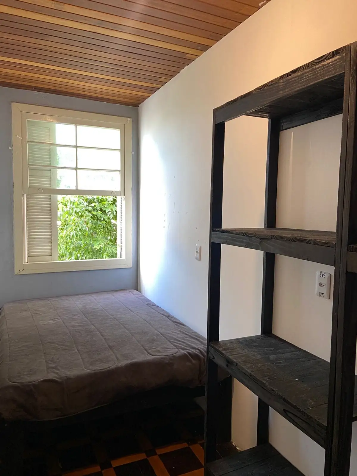 Quarto com cama, estante preta de madeira, janela com venezianas aberta para árvores e forro de madeira.