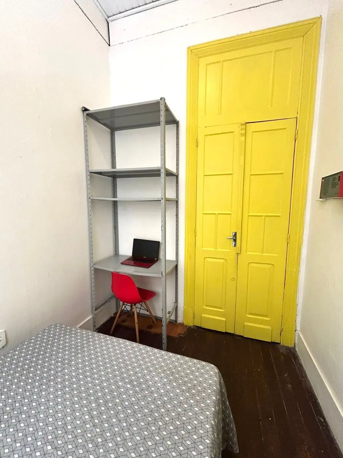Quarto com cama de lençol cinza estampado, estante de metal com notebook, cadeira vermelha e porta alta pintada de amarelo.