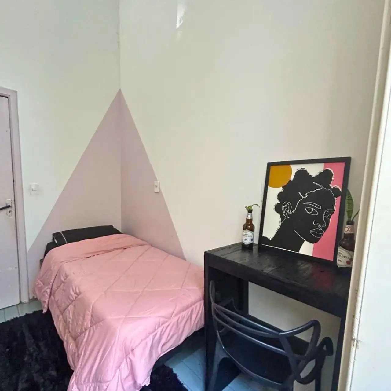 Quarto com cama de solteiro de colcha rosa, parede com triângulo rosa, mesa preta com cadeira e um quadro de ilustração apoiado sobre a mesa.