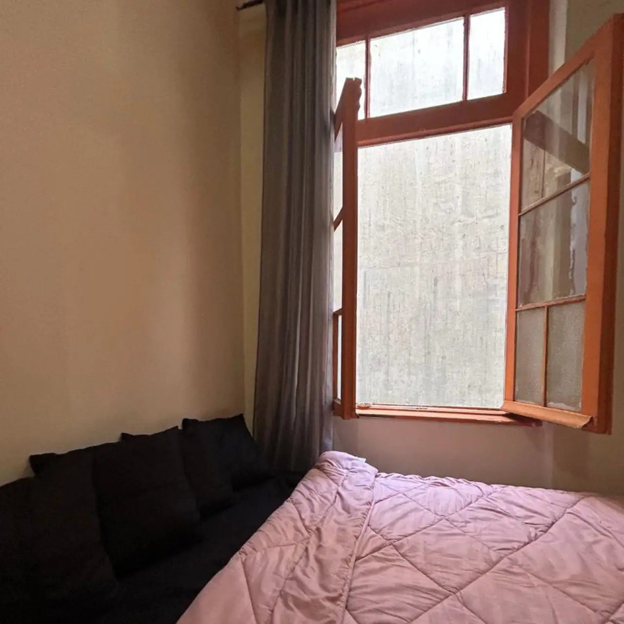 Quarto com cama de colcha rosa e almofadas pretas, janela alta de madeira laranja aberta e cortina cinza.