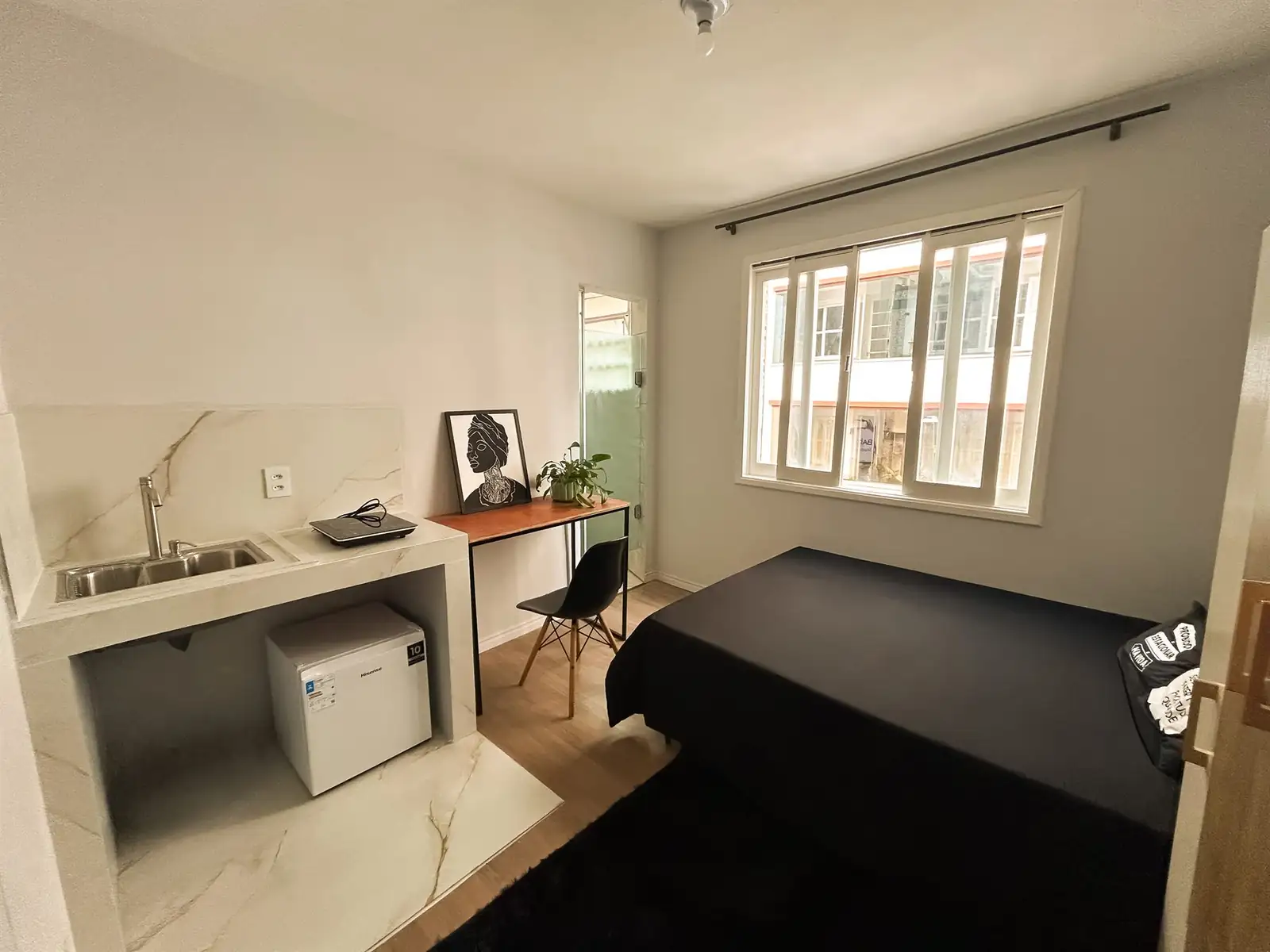 Estúdio do Bah Studios visto da porta: cozinha com pia e frigobar, mesa de trabalho, porta do banheiro e cama de casal junto à janela